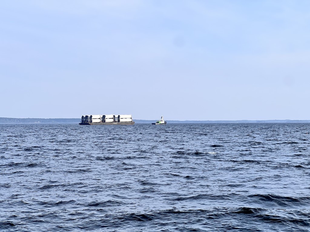 Above is a tugboat pulling barge holding wind turbine parts.